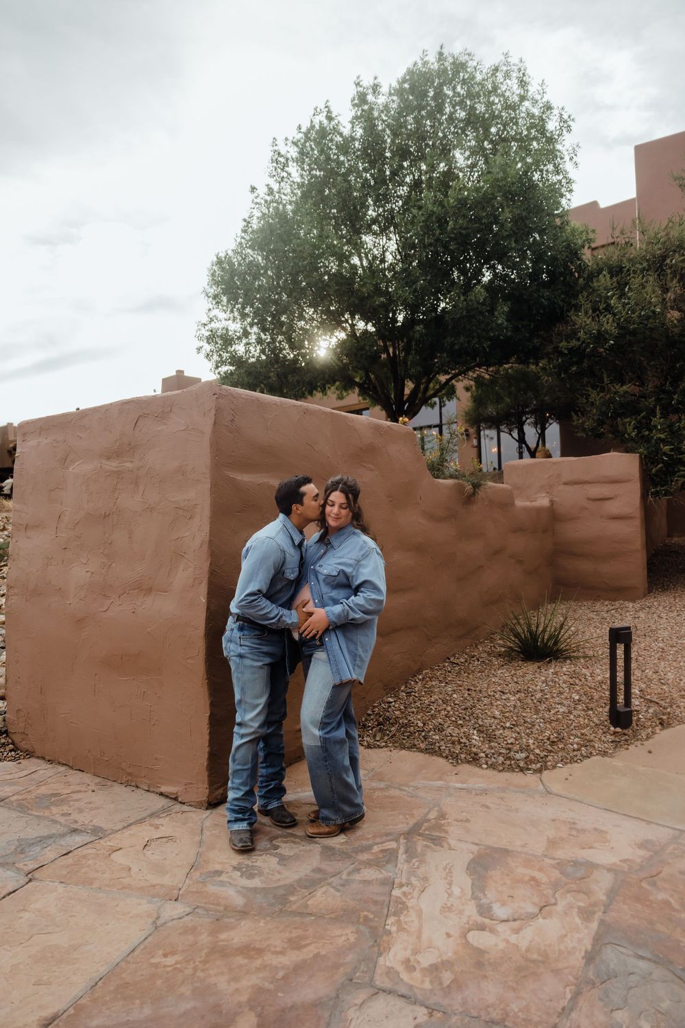 Pregnant couple sharing a tender kiss beside a terracotta adobe wall at sunset in a desert landscape.