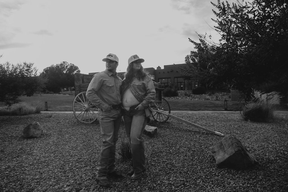 Black and white photo of expecting couple posing with vintage wagon and shovel outdoors.