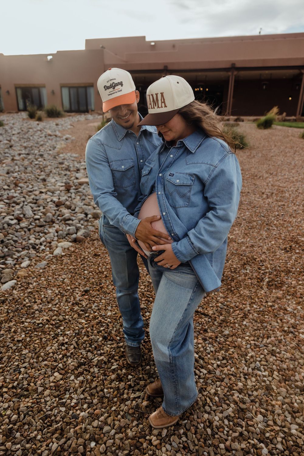 Expecting couple in matching denim and trucker hats looking down at pregnant belly on a gravel path.