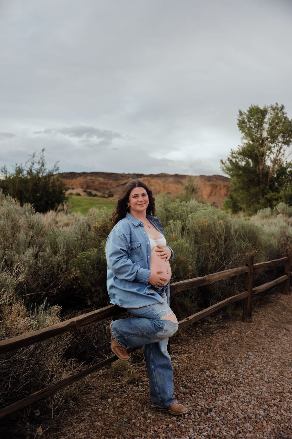 Pregnant woman in denim shirt leans on wooden fence in desert landscape holding baby bump on cloudy day.
