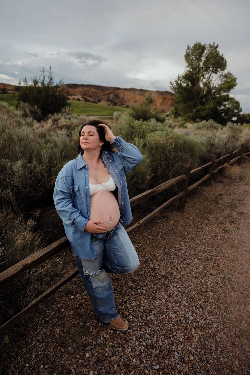Pregnant woman in denim shirt cradles baby bump while leaning on rustic fence in sagebrush landscape.