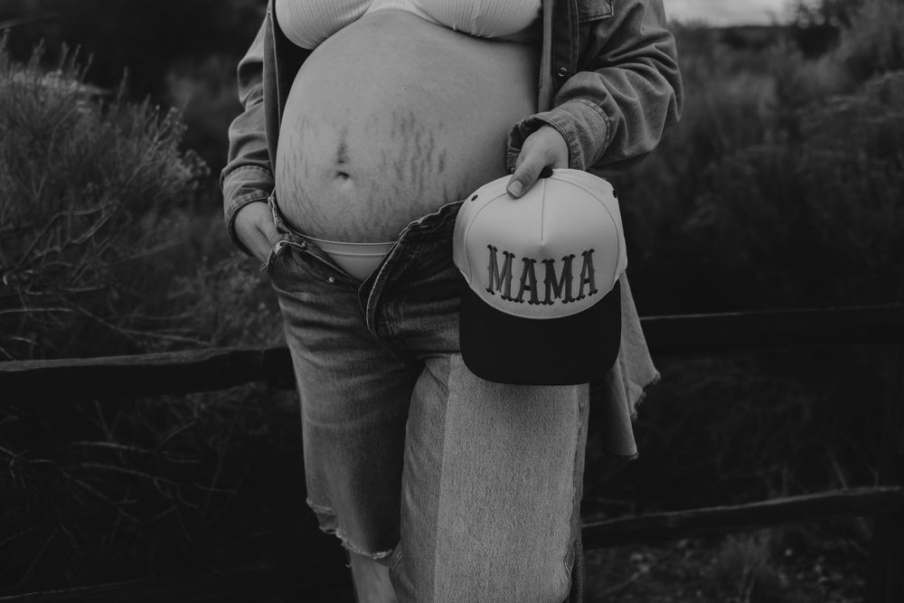 Black and white close-up of pregnant woman holding MAMA trucker hat beside exposed baby bump outdoors.