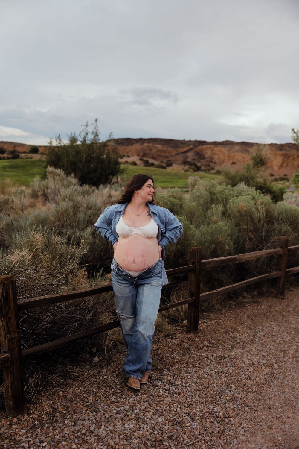 Pregnant woman in open denim jacket and white bralette leans on wooden fence in desert shrubland setting.