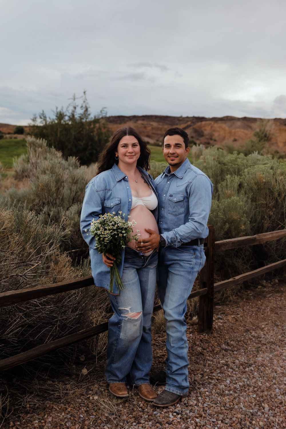 Expecting couple in matching denim outfits pose together holding wildflowers by rustic fence in desert.