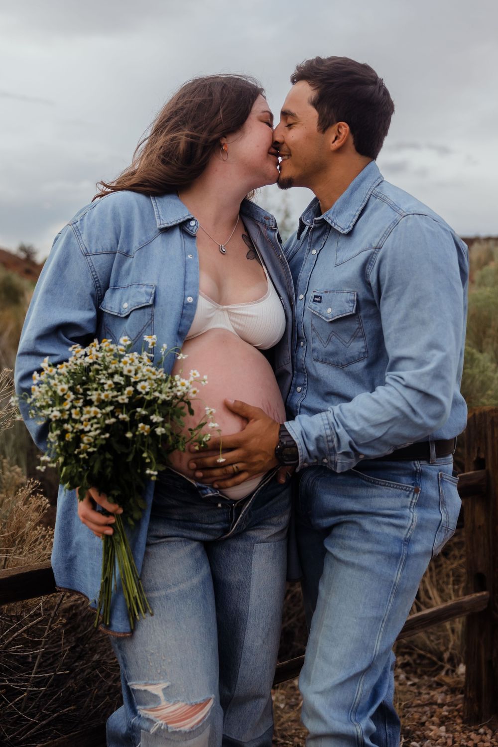 Expecting couple in denim outfits share a kiss while man cradles baby bump holding wildflower bouquet.