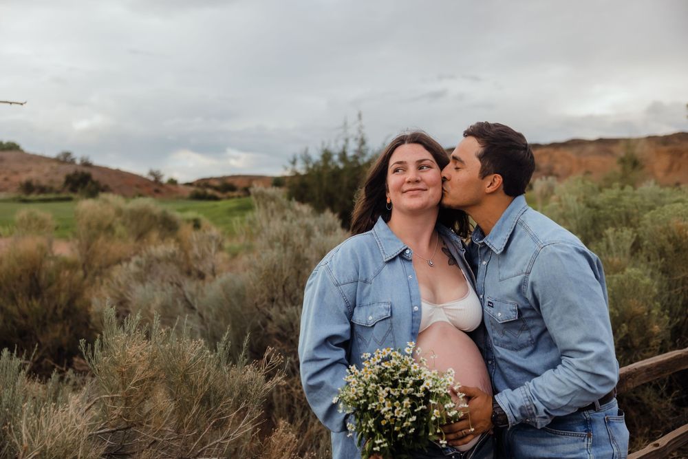 Pregnant couple in matching denim embracing outdoors, man kissing woman's cheek as she holds wildflower bouquet.