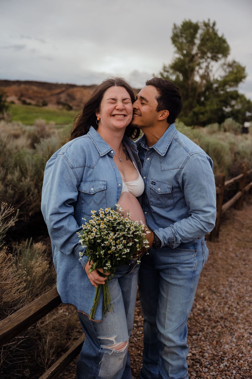 Joyful pregnant couple in denim outfits laughing together outdoors, woman holding bouquet of white wildflowers.