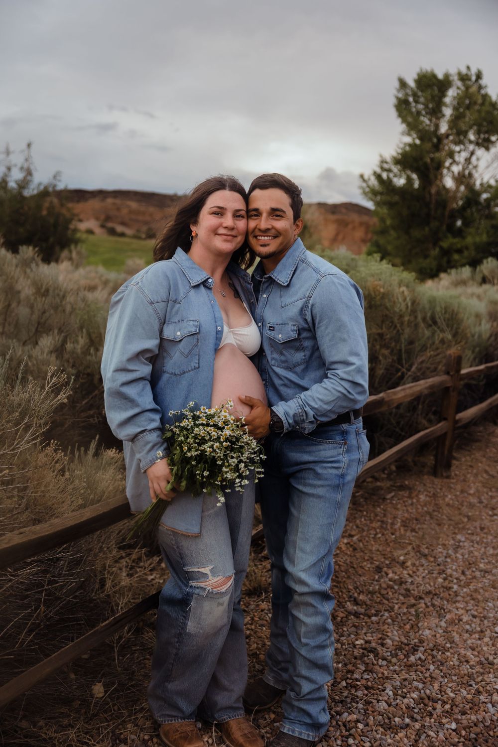 Smiling pregnant couple in coordinating denim outfits posing together outdoors, woman holding white wildflower bouquet.