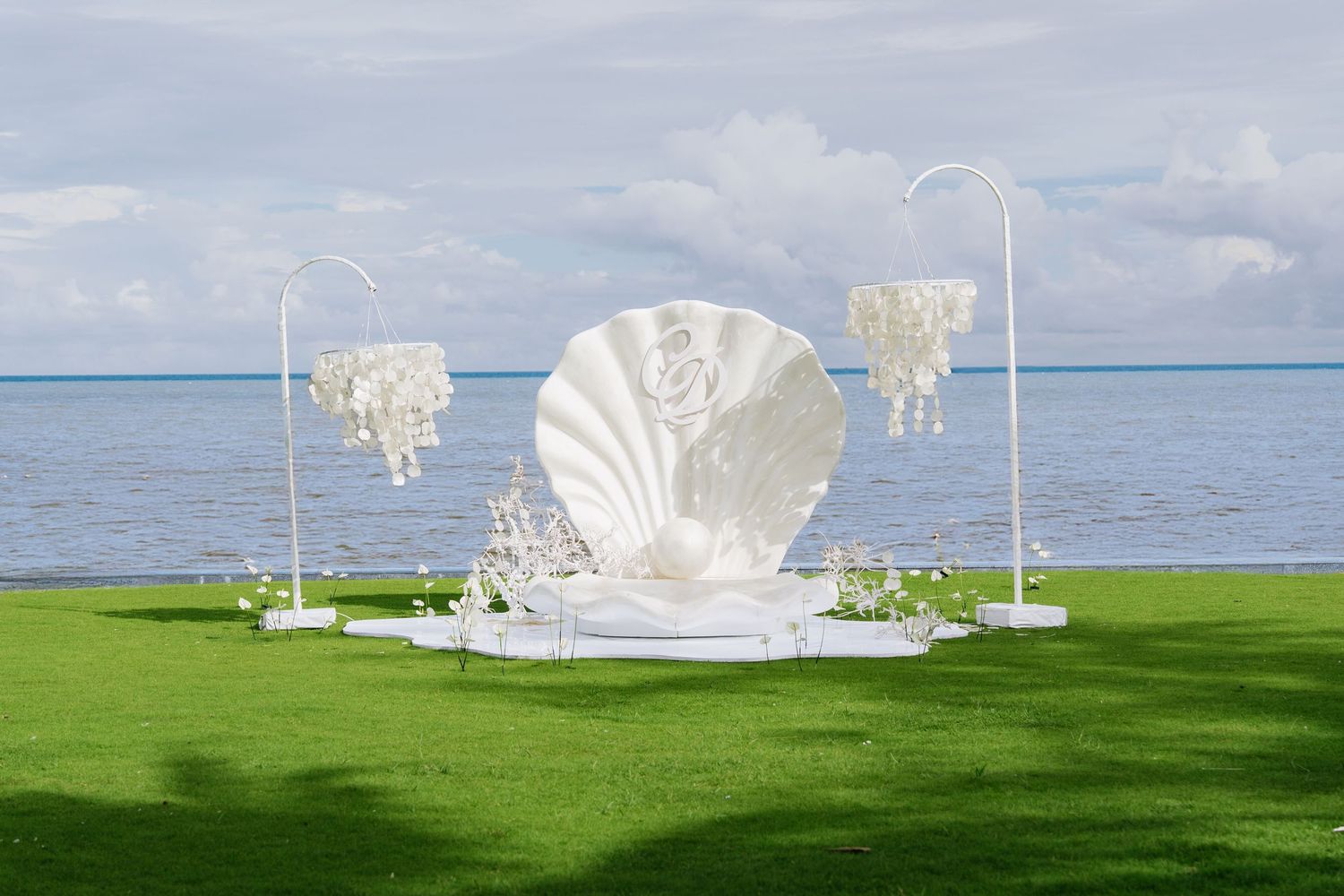 White giant seashell wedding altar with pearl chandeliers on green lawn by the sea.
