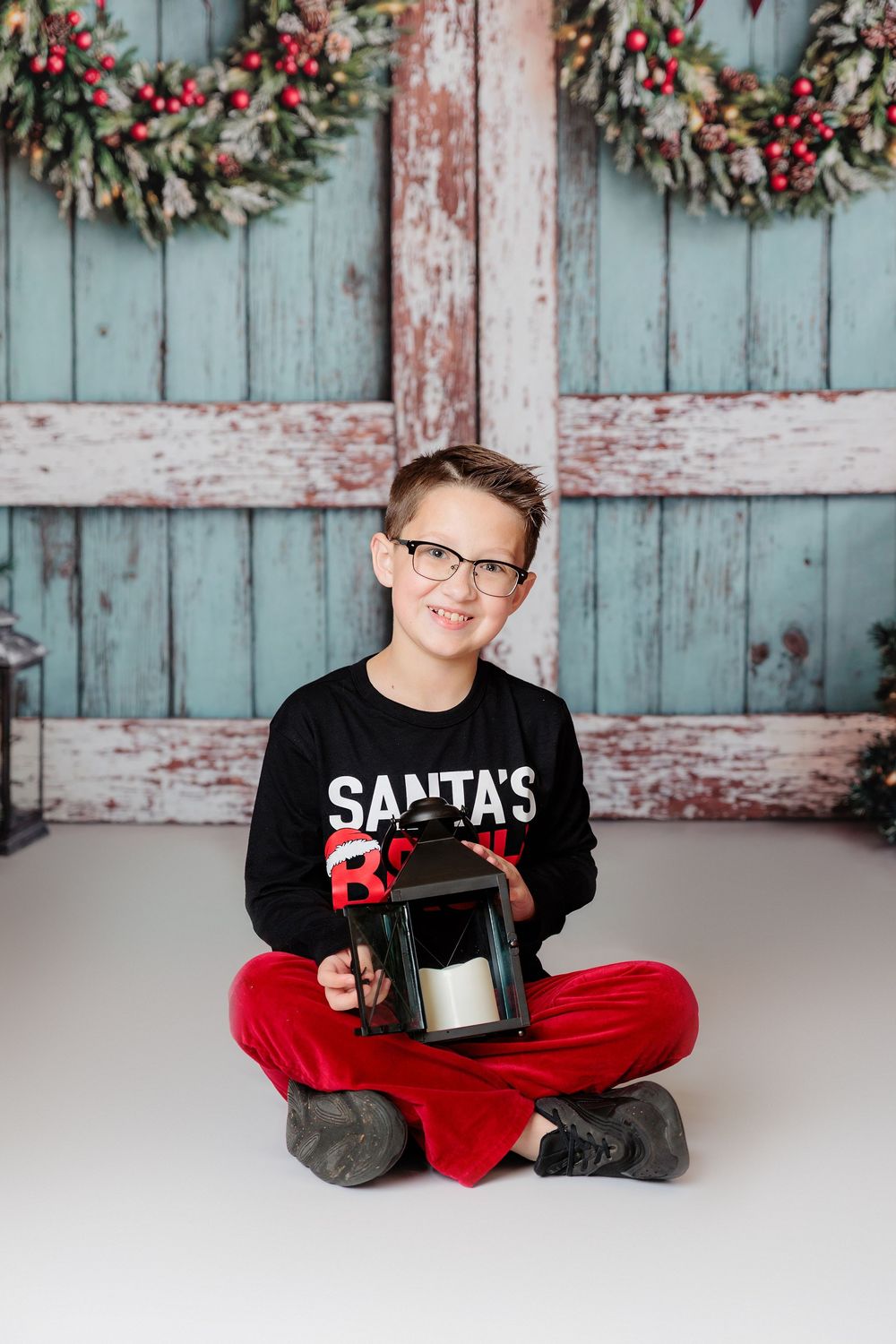 Cheerful person in Christmas themed sleepwear poses cross-legged against weathered blue barn door with wreaths studio session clayton nj