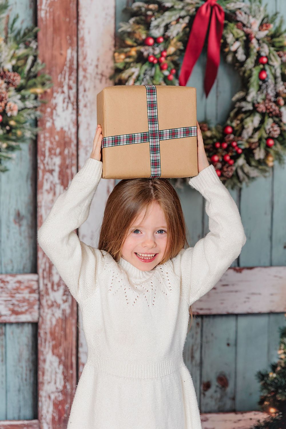 Child in white sweater holds wrapped Christmas present in front of rustic blue door with holiday wreath and decor. studio session clayton nj