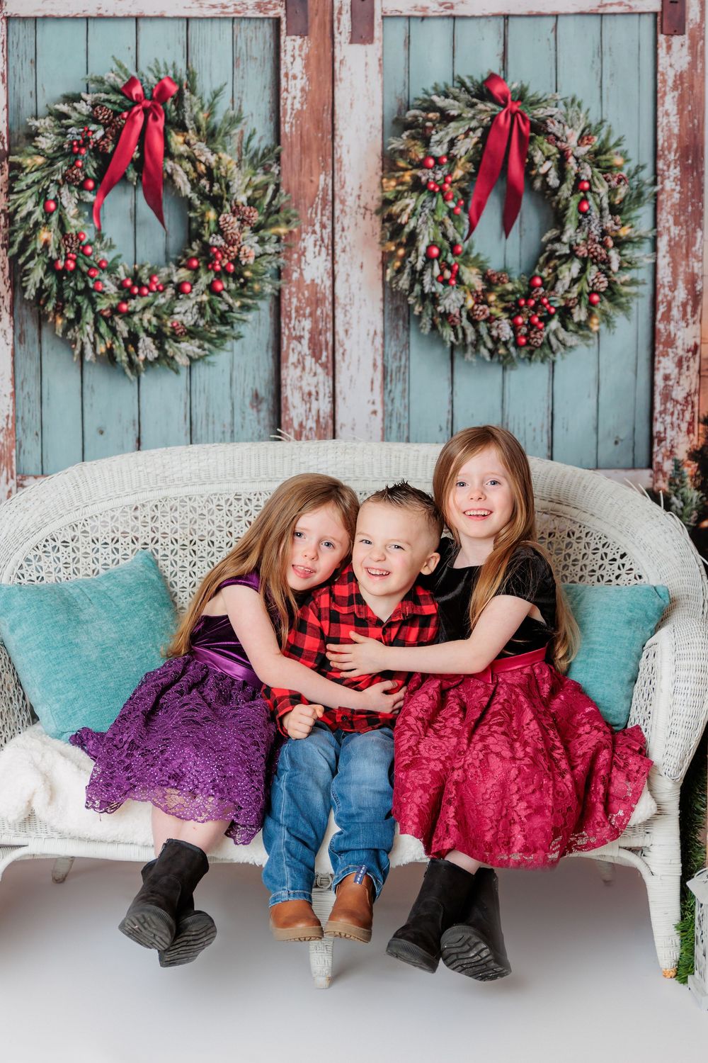 Three children pose on a wicker bench against rustic blue doors decorated with festive Christmas wreaths. studio session clayton nj