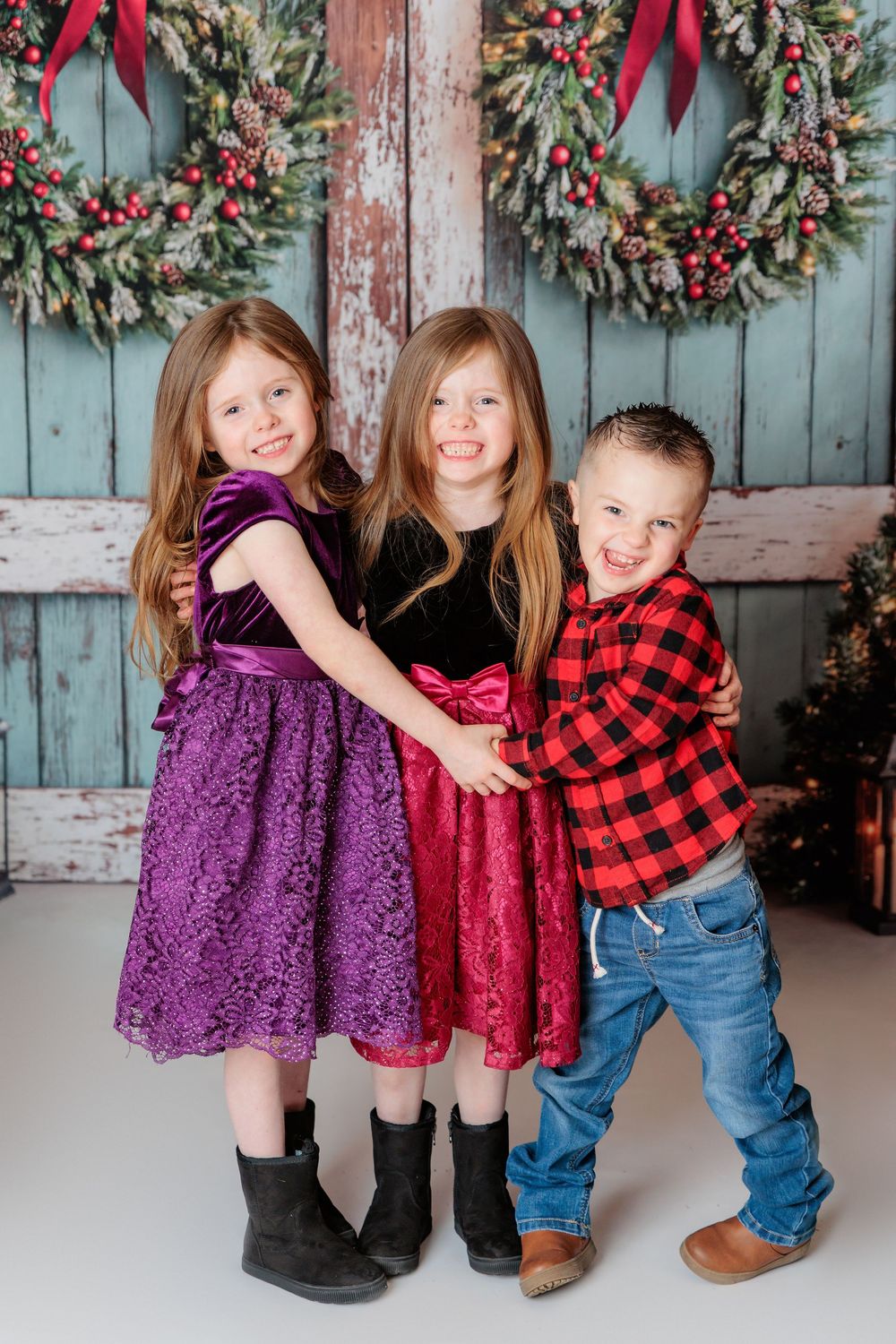 Children dressed in festive holiday outfits stand against a rustic blue wall decorated with a Christmas wreath. studio session clayton nj