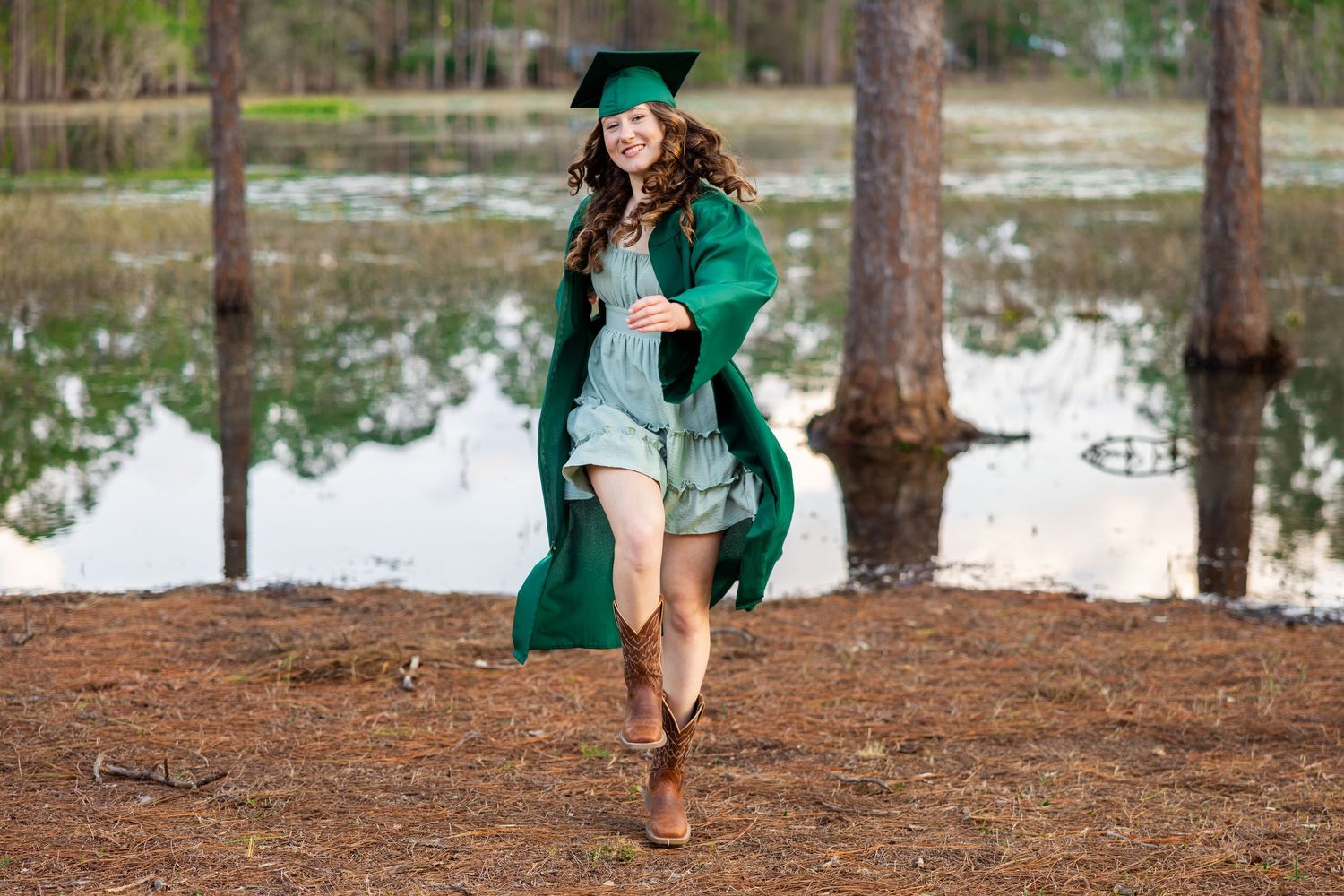 Woman in turquoise graduation gown walks through pine trees near a lake during outdoor graduation photo session.