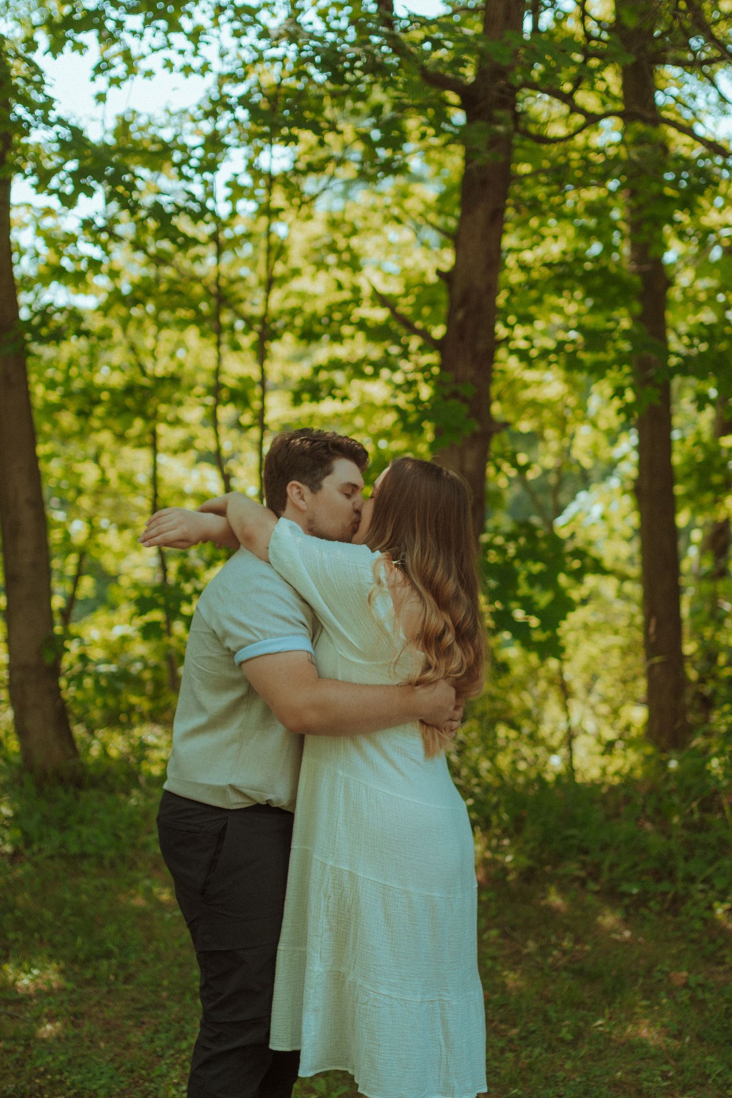A romantic photoshoot in a sunlit forest setting shows a couple embracing among green trees and foliage.