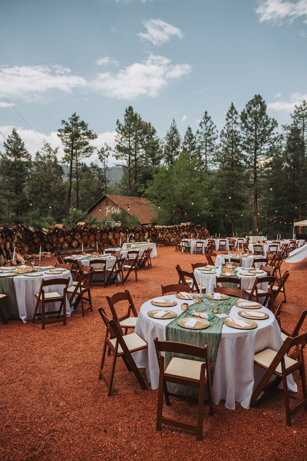 Outdoor wedding reception setup with round tables and wooden chairs on red dirt ground surrounded by pine trees.