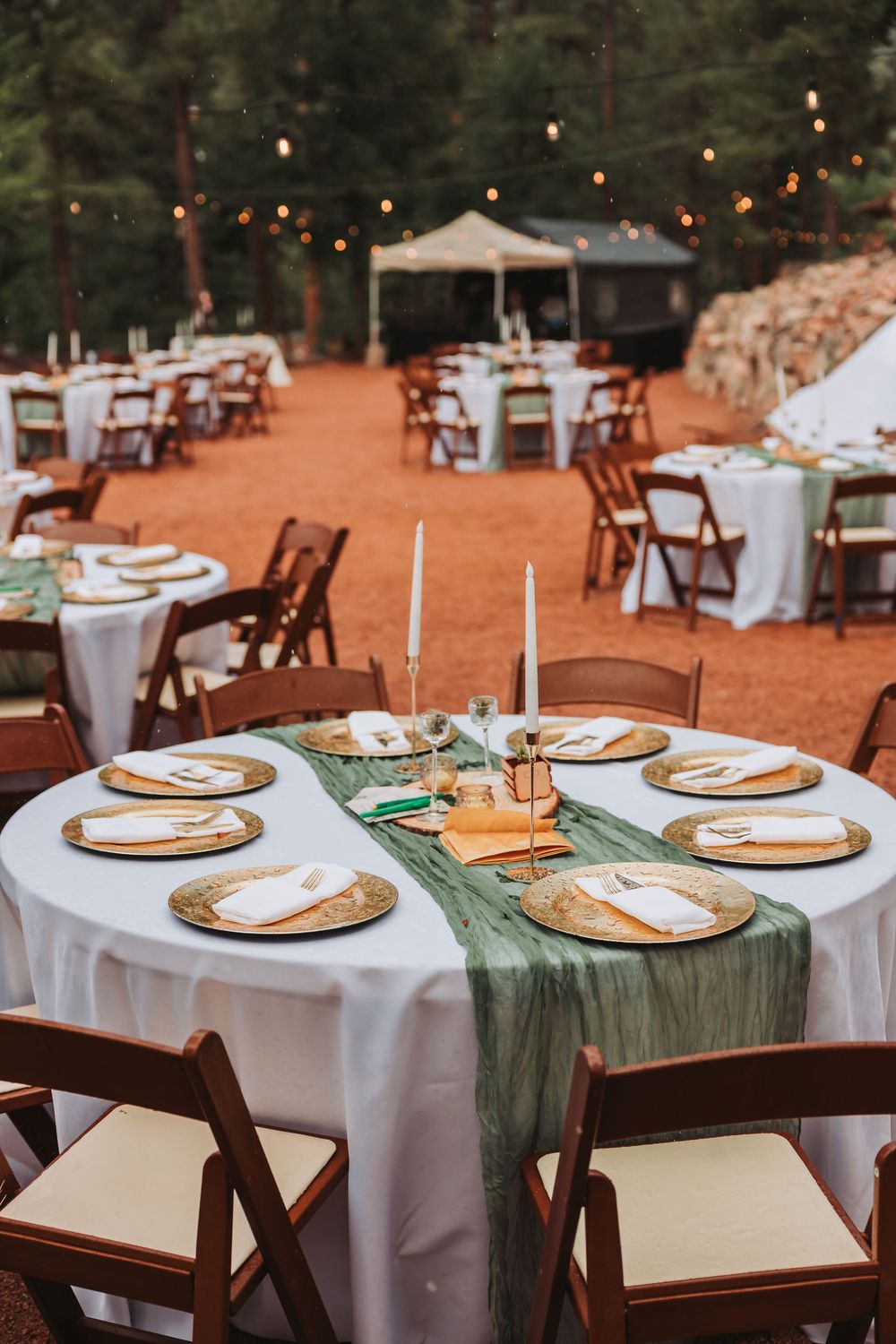 Outdoor wedding reception tables set with white linens, rustic charger plates, and sage green table runners on red clay ground.