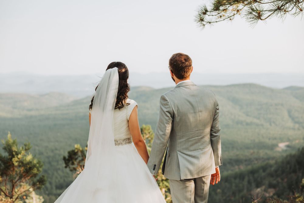 Bride and groom standing on mountain overlook admiring scenic forest vista during outdoor wedding ceremony.