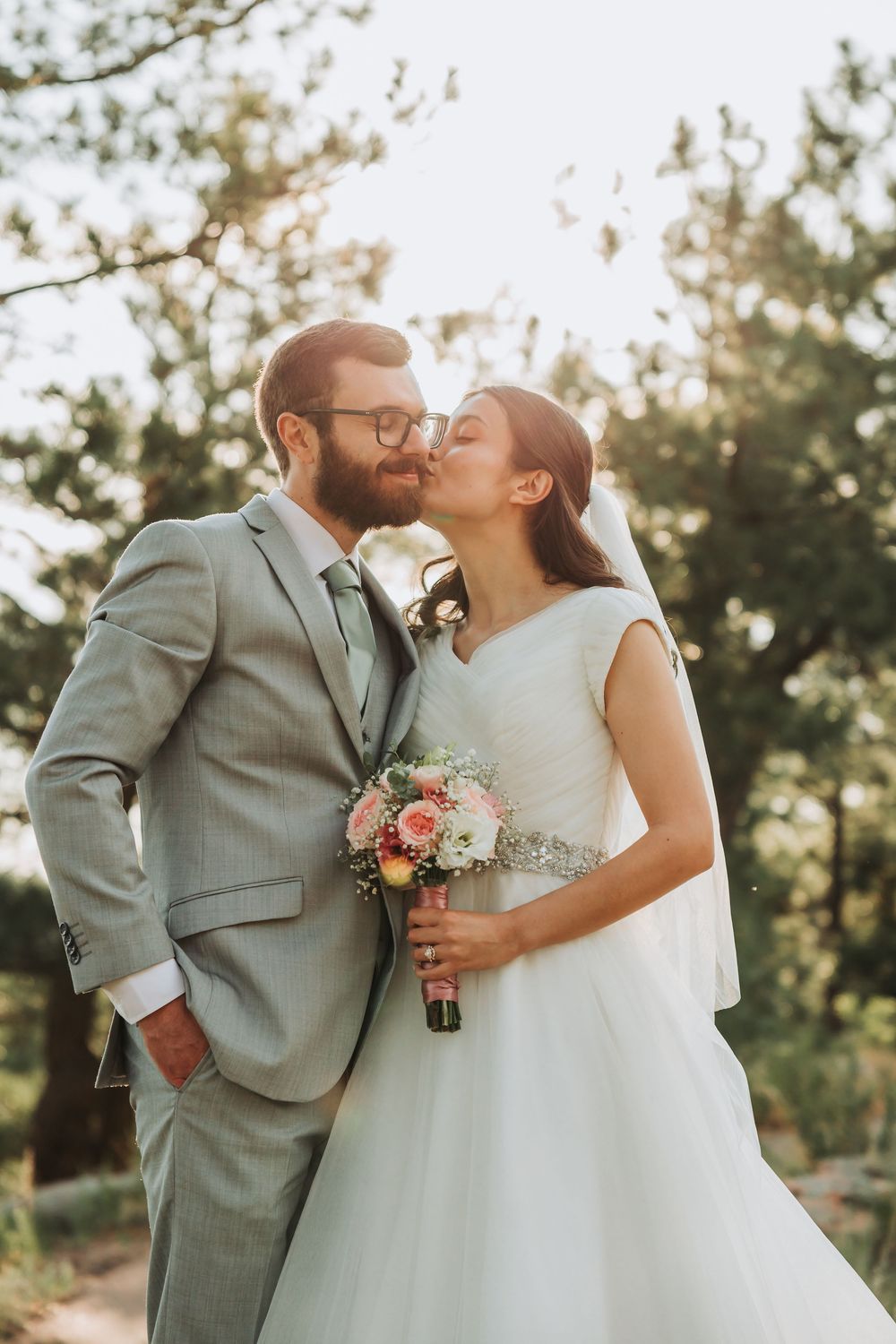 A romantic wedding couple shares three sunset kisses while holding a delicate bouquet in a dreamy outdoor setting with golden light.