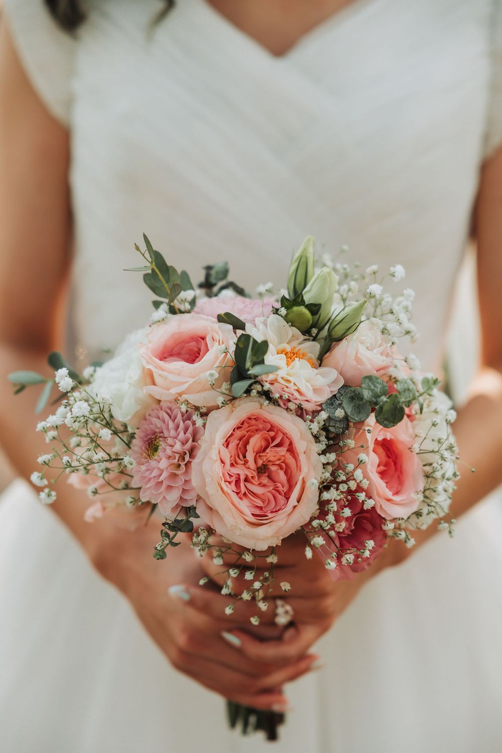 Four delicate pink garden rose wedding bouquets with baby's breath and greenery held by hands in white dresses.