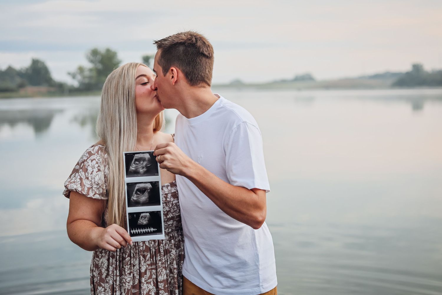 Couple holding ultrasound photo by serene waterfront during sunset announcement session. Announcement photography