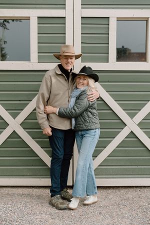 A couple in casual western wear embraces against a green barn door with white trim.