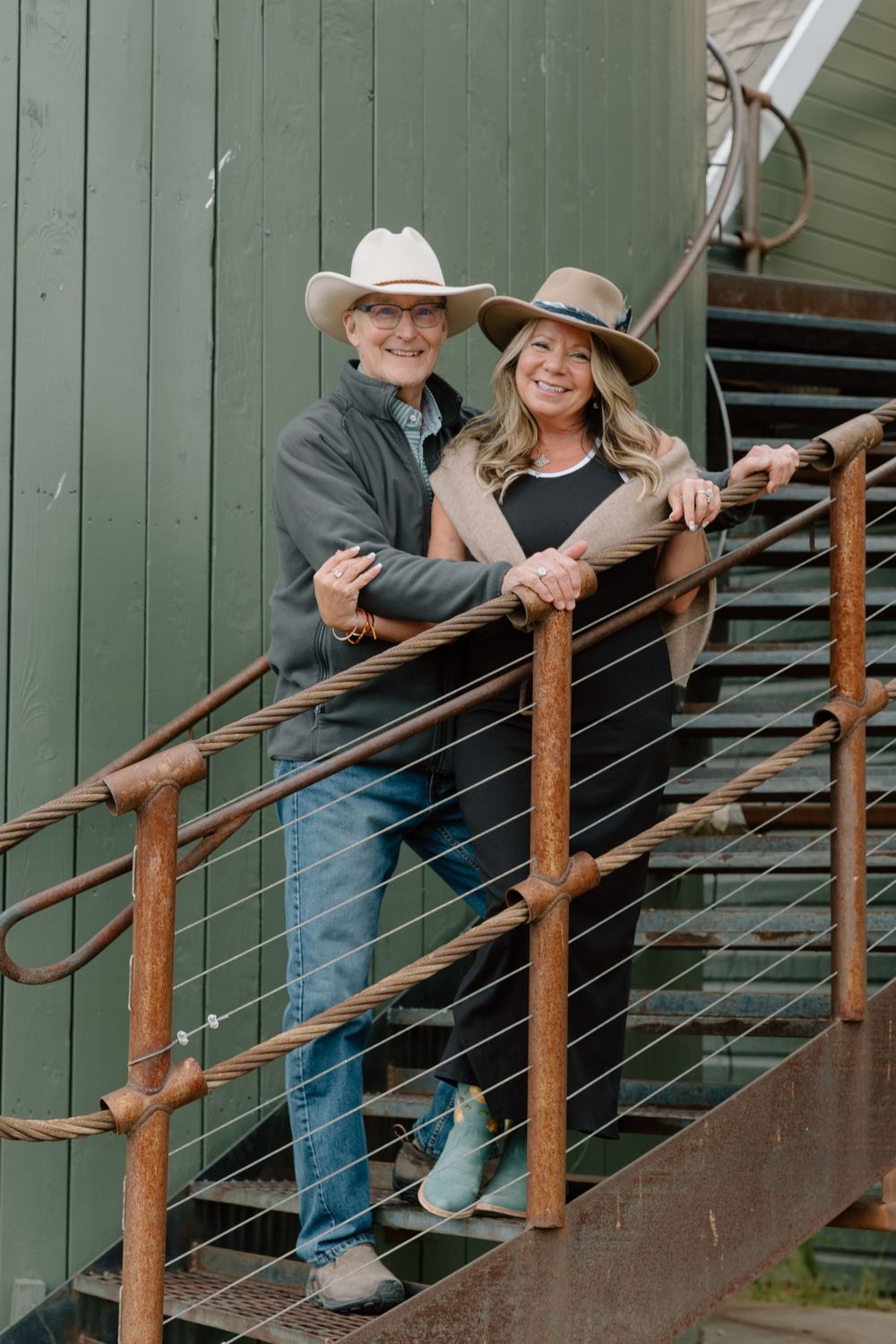 A couple wearing cowboy hats stands together on rustic wooden stairs by a green building.