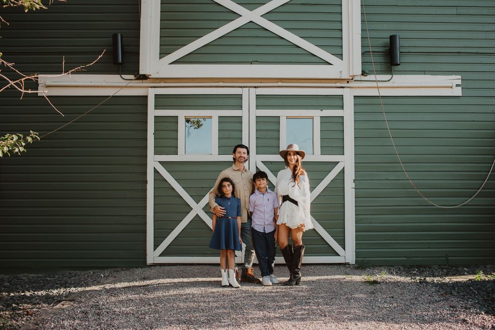 A family stands in front of a large green barn with white trim and decorative X patterns.
