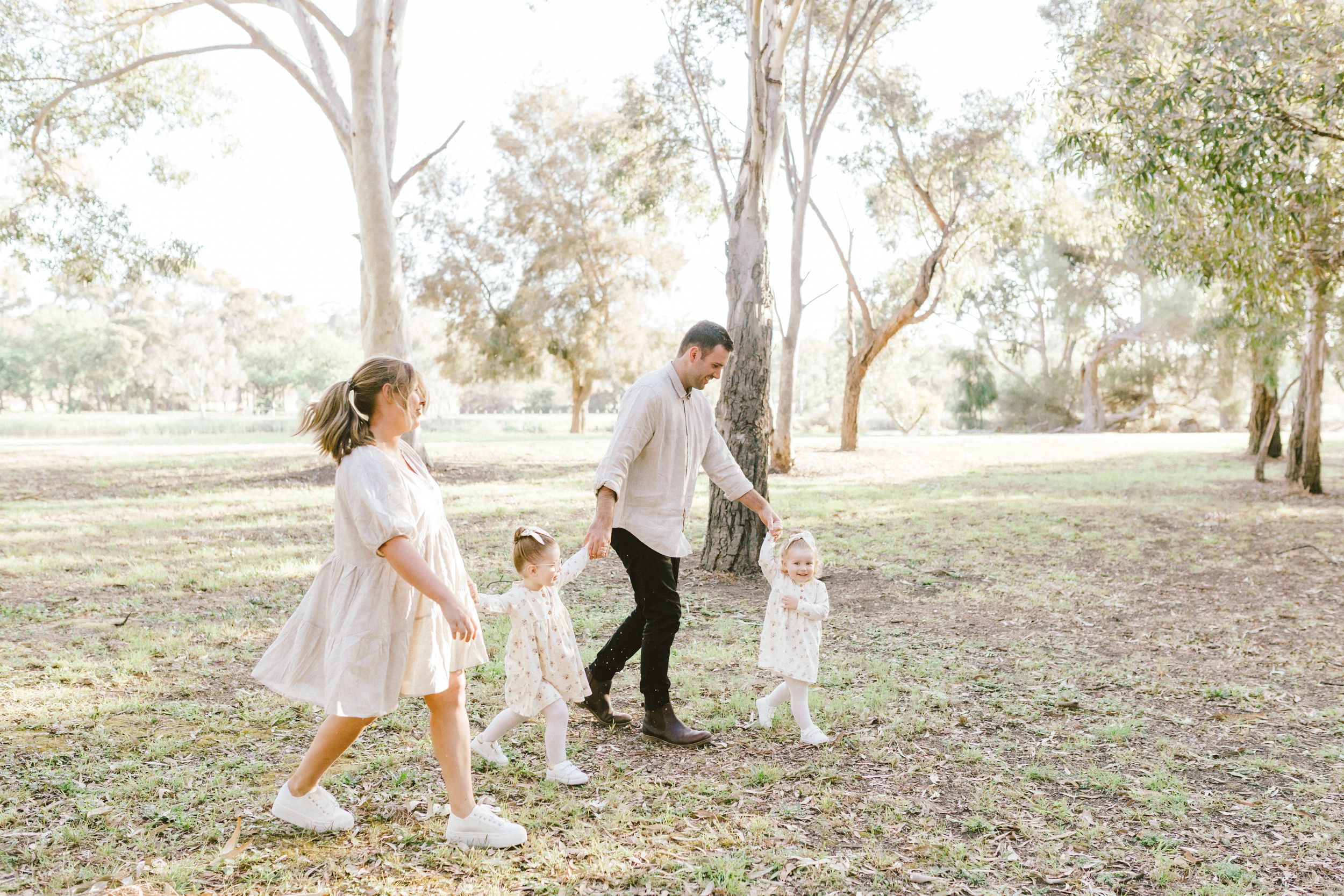 Family wearing white clothing plays together on a grassy hillside under eucalyptus trees on a bright sunny day.