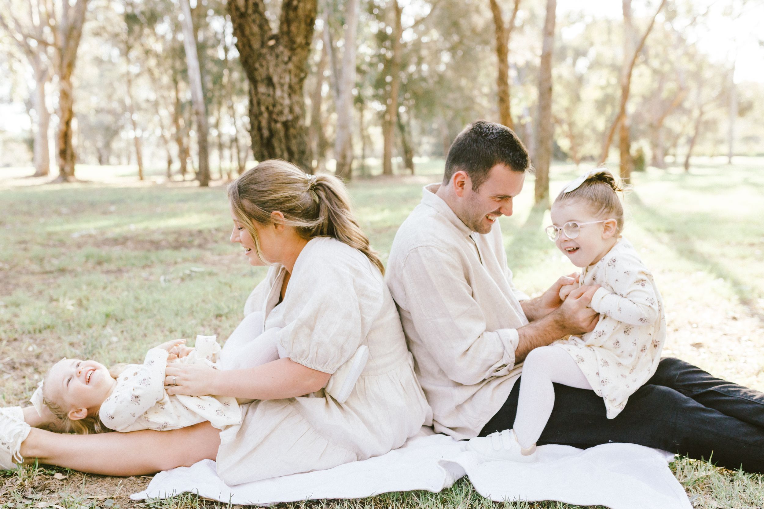 Family members in white outfits lie together on grass during an outdoor photo session in natural lighting.