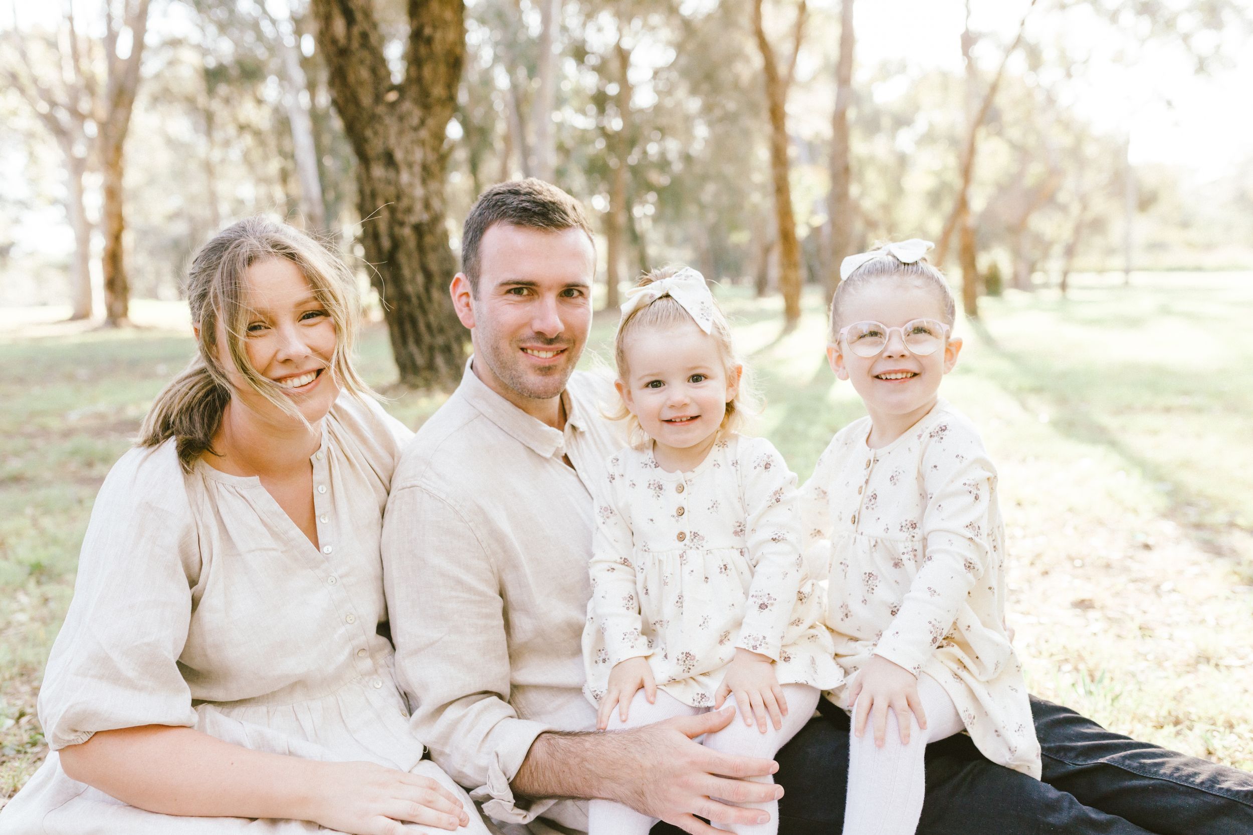 A family dressed in white and cream colored outfits sits together outdoors in a sunlit wooded setting.