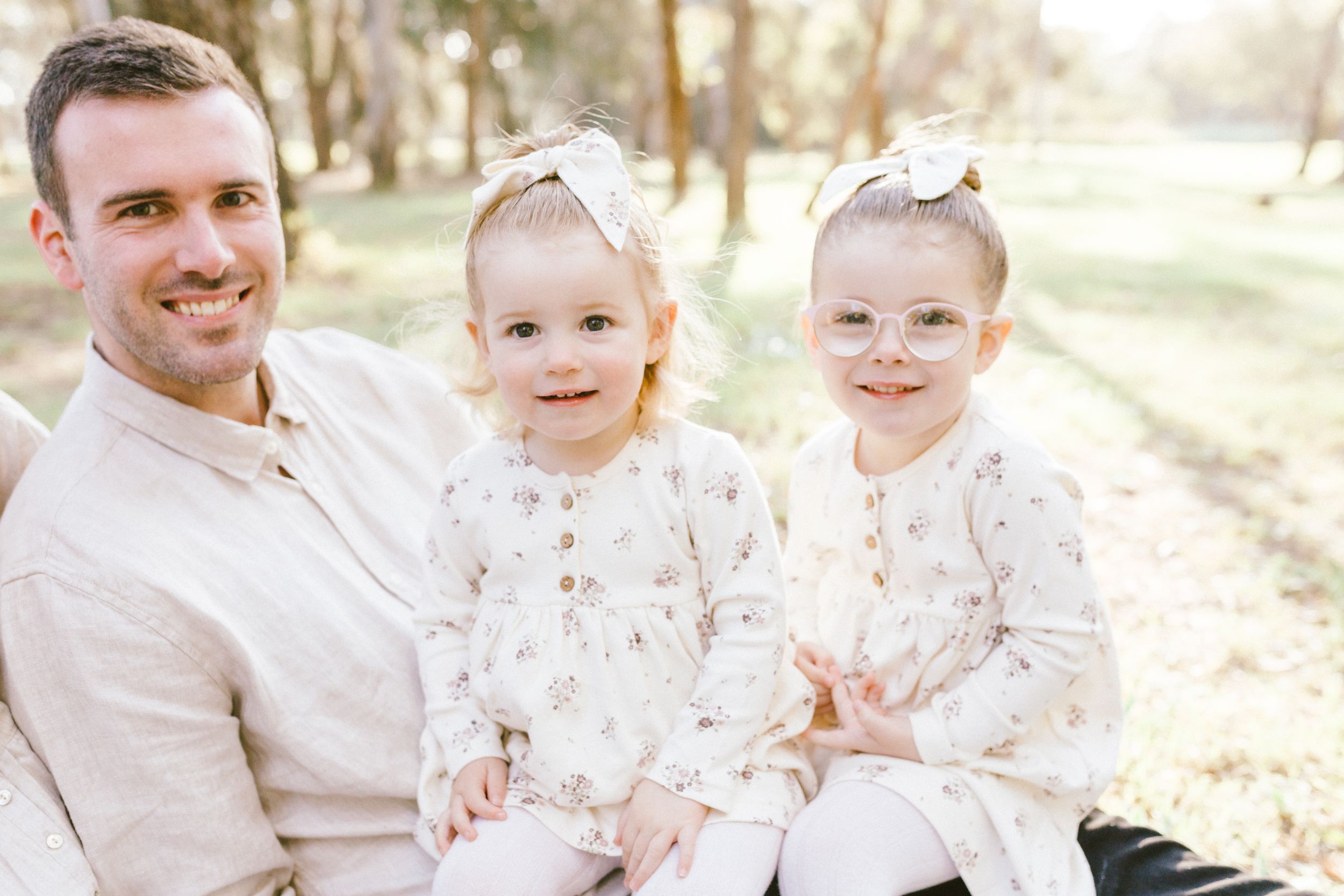 A family group dressed in white and beige clothing sits together outdoors in warm sunlight during a photo session.