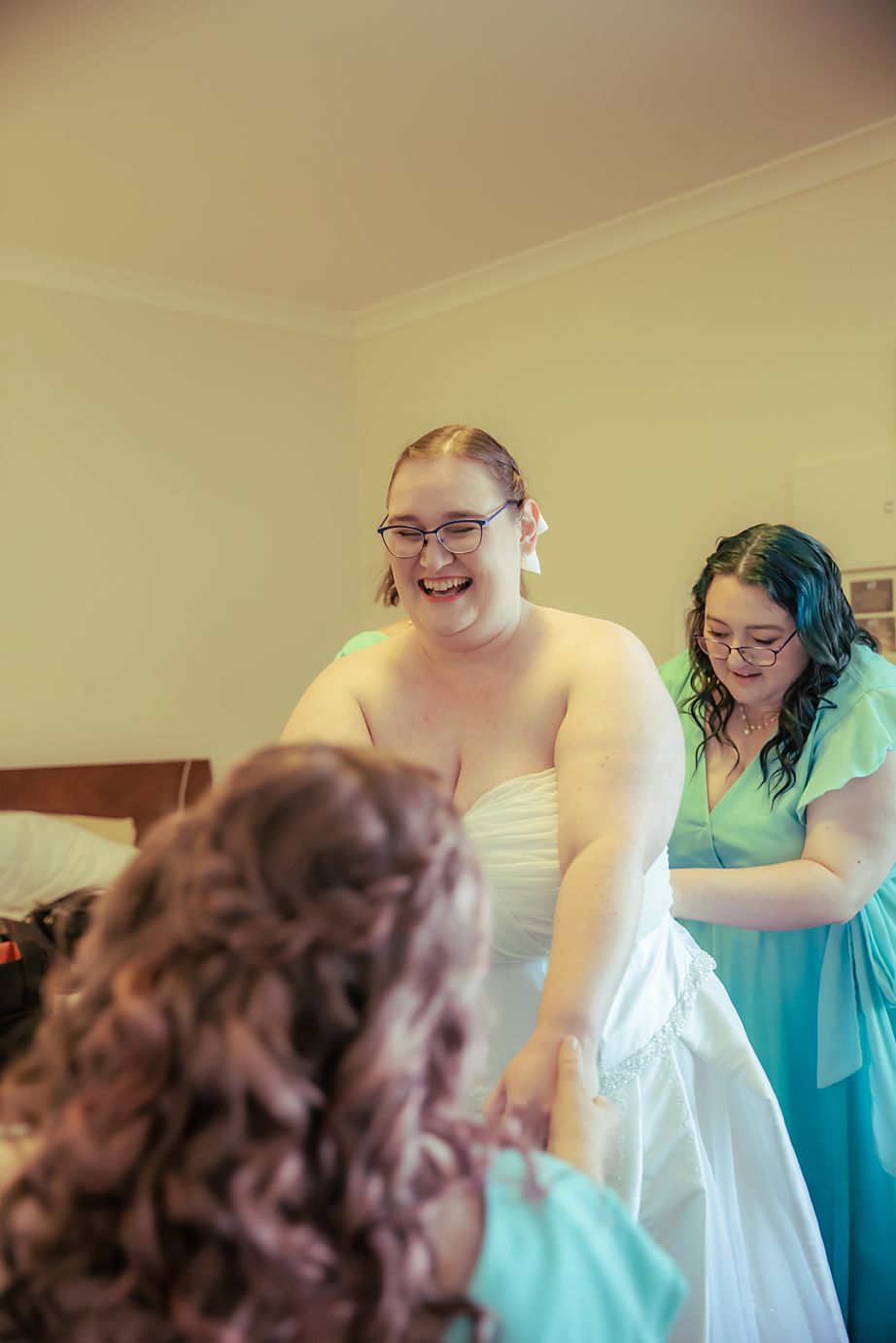 A joyful person in a wedding dress gets help from attendants wearing turquoise during wedding preparations.