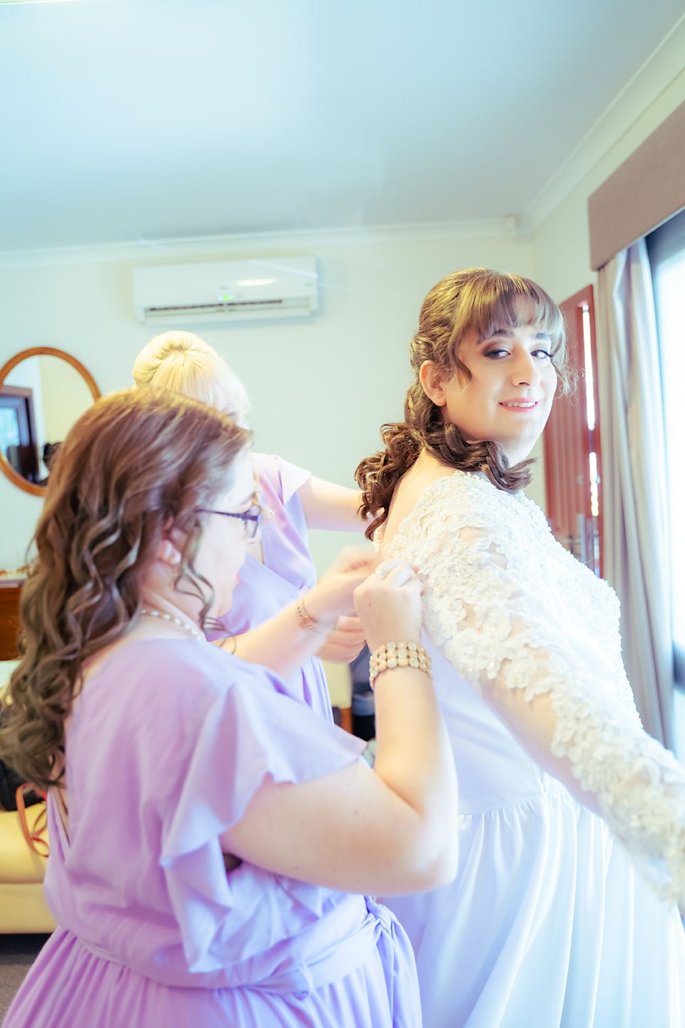 Several bridesmaids in light purple dresses help adjust a white wedding dress during preparations.
