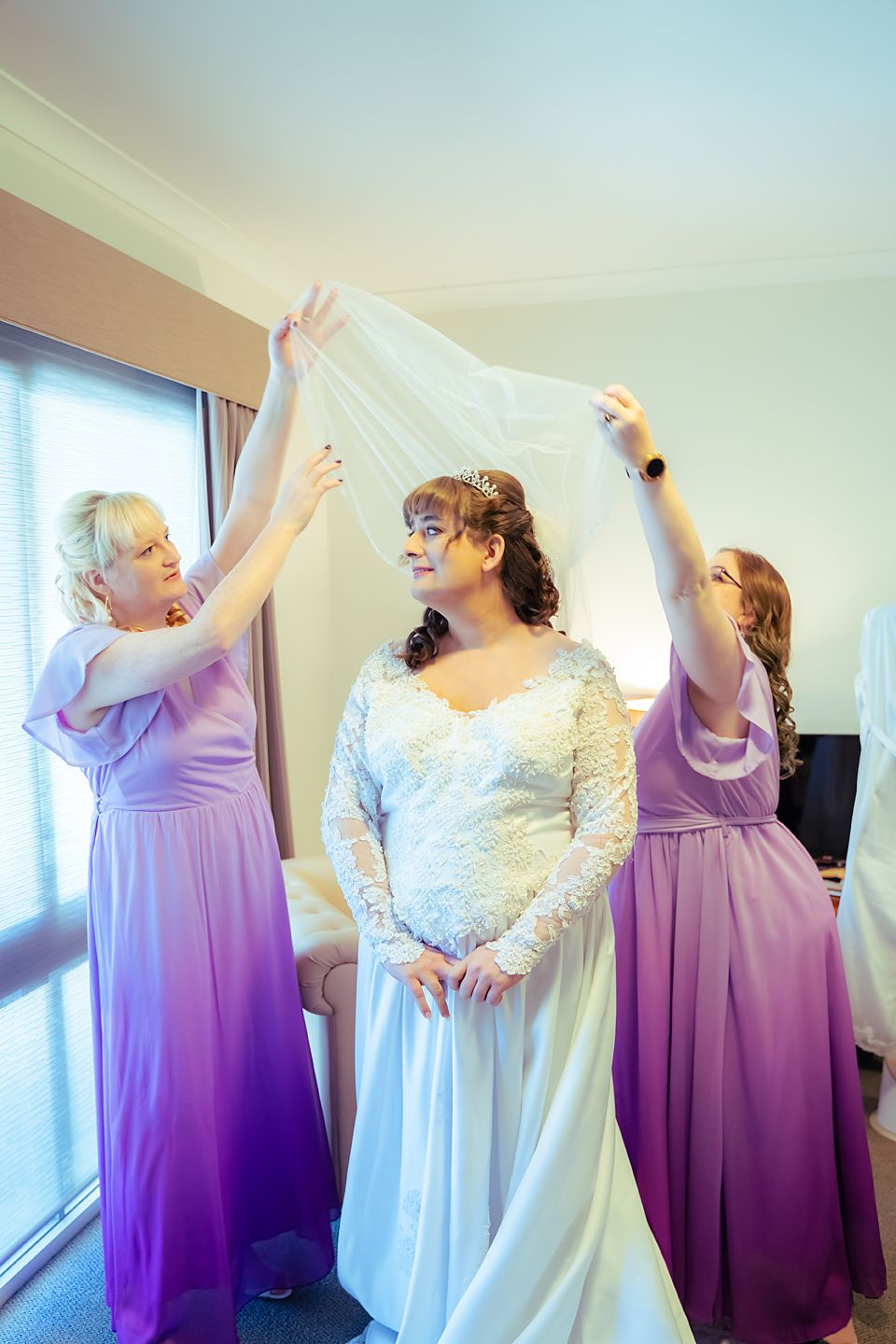 Bridesmaids in lavender dresses assist with the bride's veil near a bright window in a wedding preparation room.