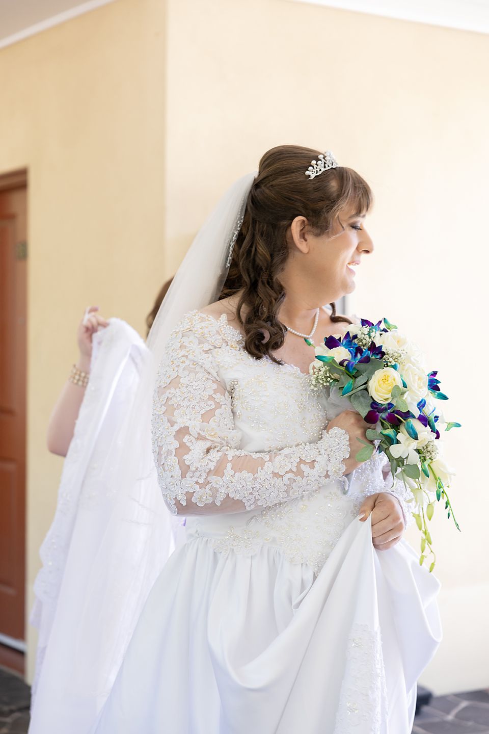 A bride in a white lace wedding dress holds a vibrant blue and yellow bouquet while posing.