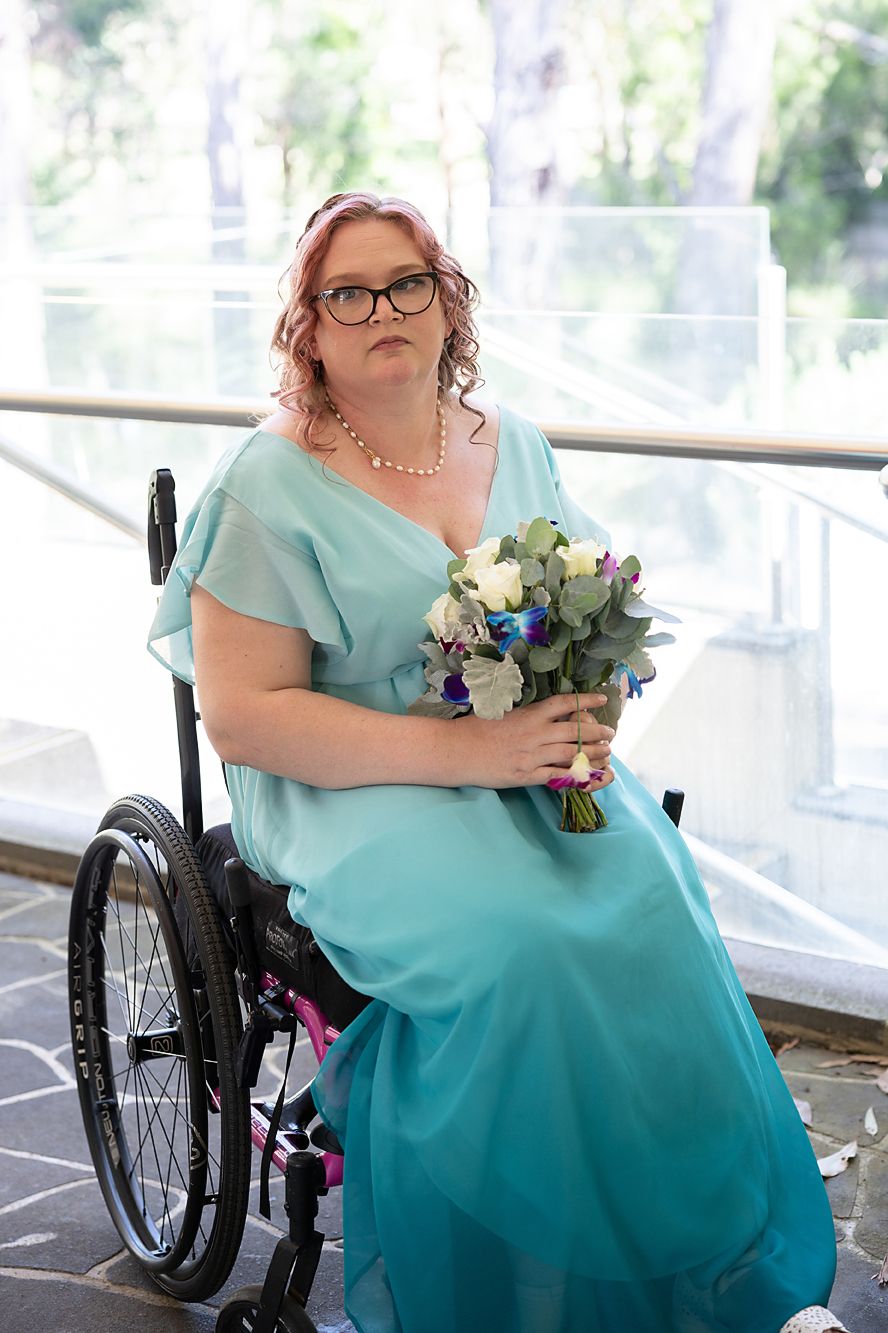A person in a teal bridesmaid dress and wheelchair holds a bouquet during a wedding celebration.