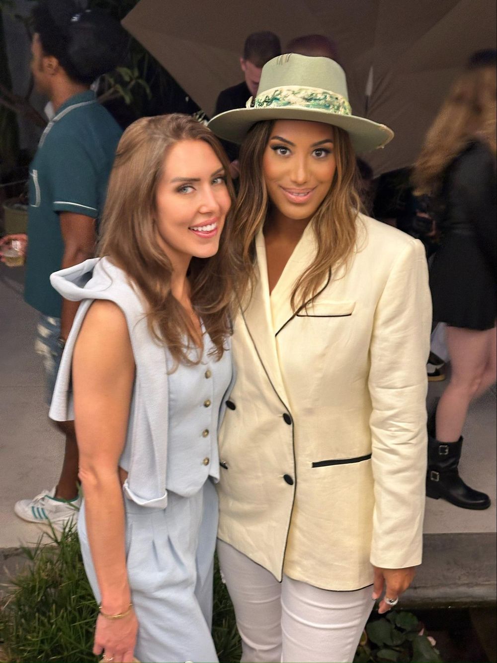 Two women pose at an event, one wearing a white blazer and hat, the other in a light-colored outfit.