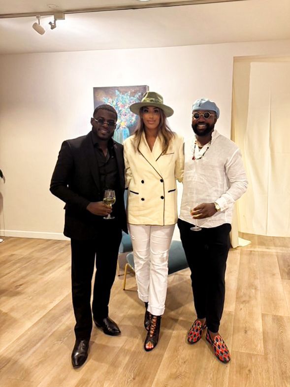 Three people dressed in stylish attire pose together in a minimalist room with wooden flooring and white walls.