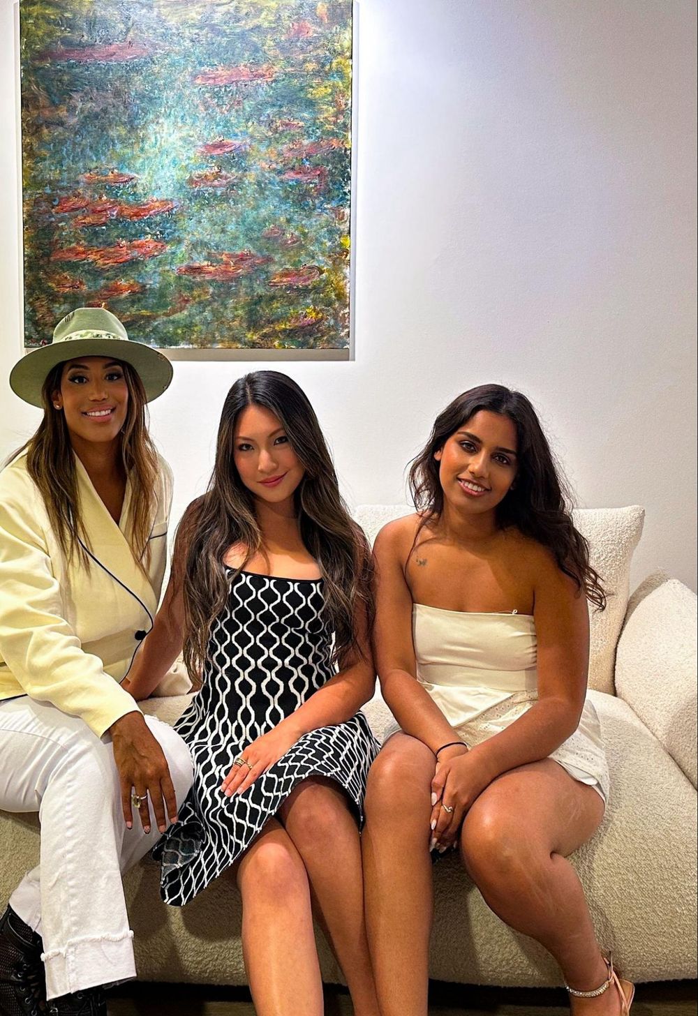 Three friends sit on a white couch beneath colorful abstract wall art, wearing stylish summer outfits in yellow and white.