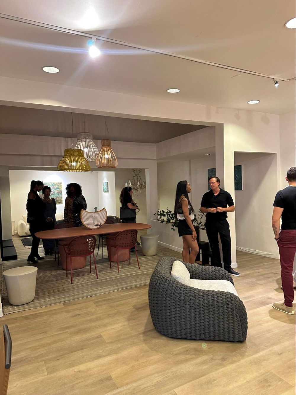 Modern office space with wood floors, recessed lighting, a wicker lounge chair and people gathering in a casual setting.