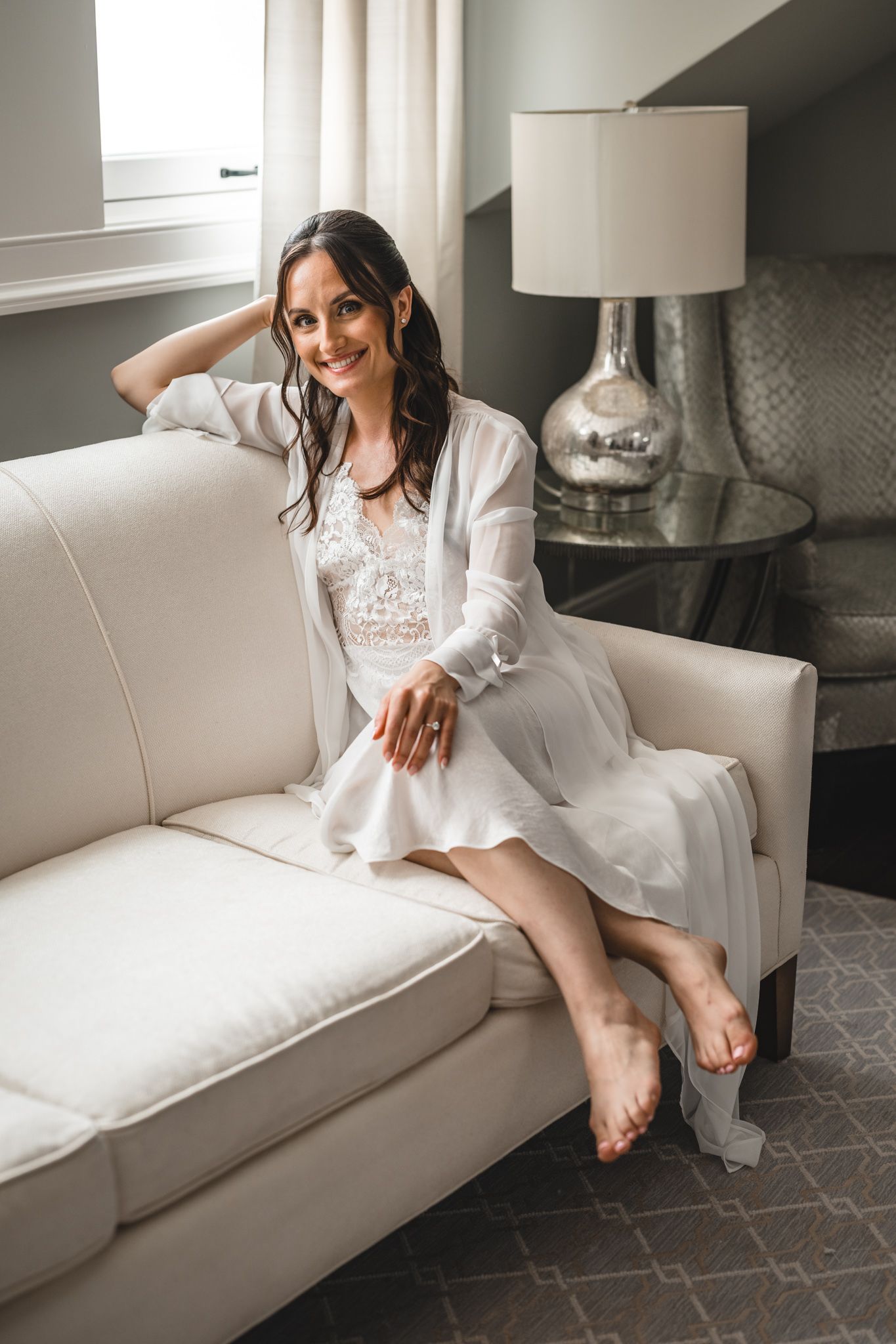 Bride in white robe relaxing on white couch during wedding preparations.