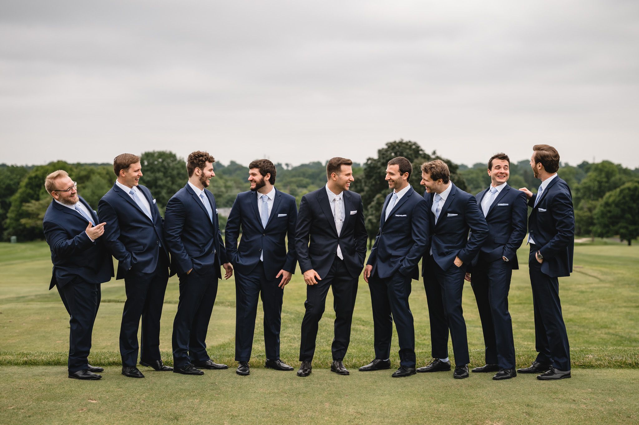 Wedding party in navy suits poses creatively on grassy field for group photos.