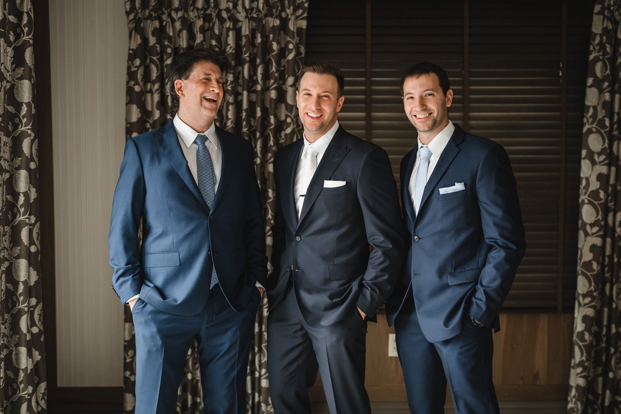 Groomsmen in matching navy suits share candid moments against floral curtain backdrop.