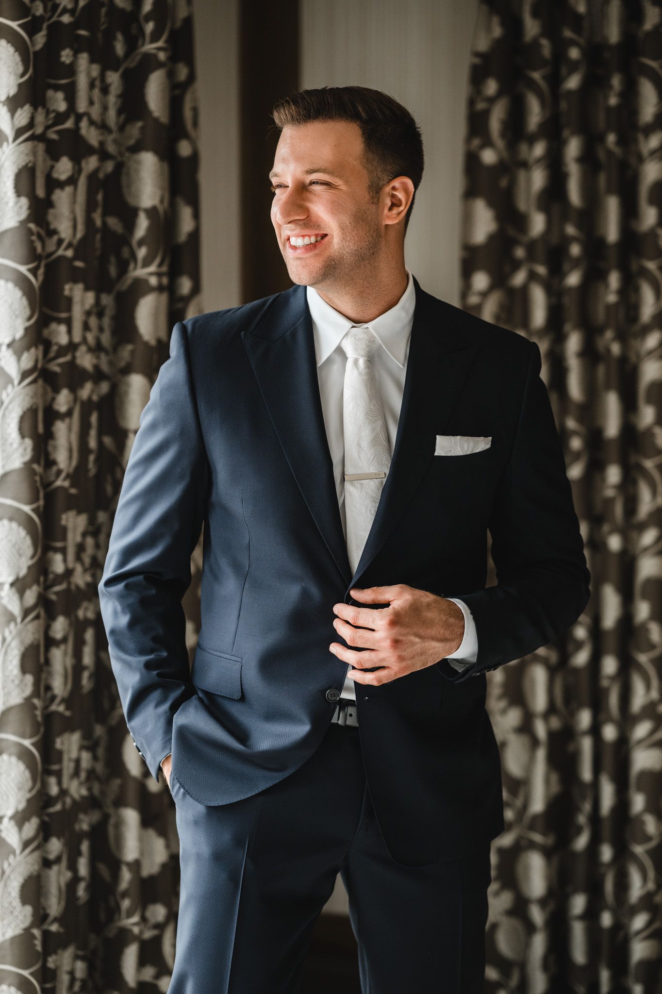 Elegant series of poses in a navy suit and white tie against floral curtains.