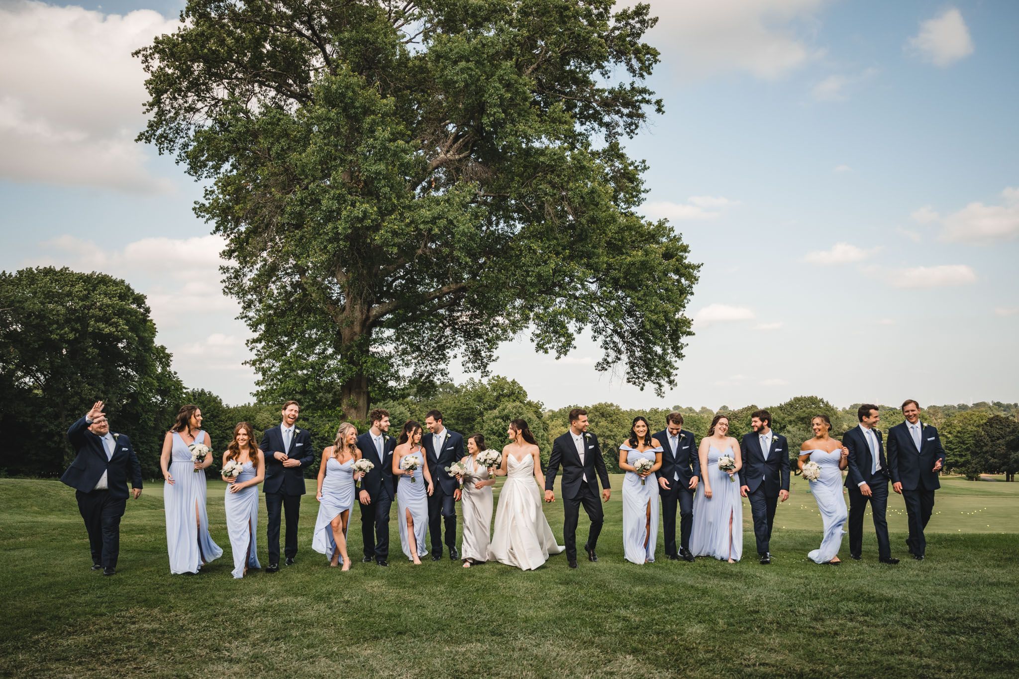 Wedding party lined up for photos on a grassy hill under blue summer sky.
