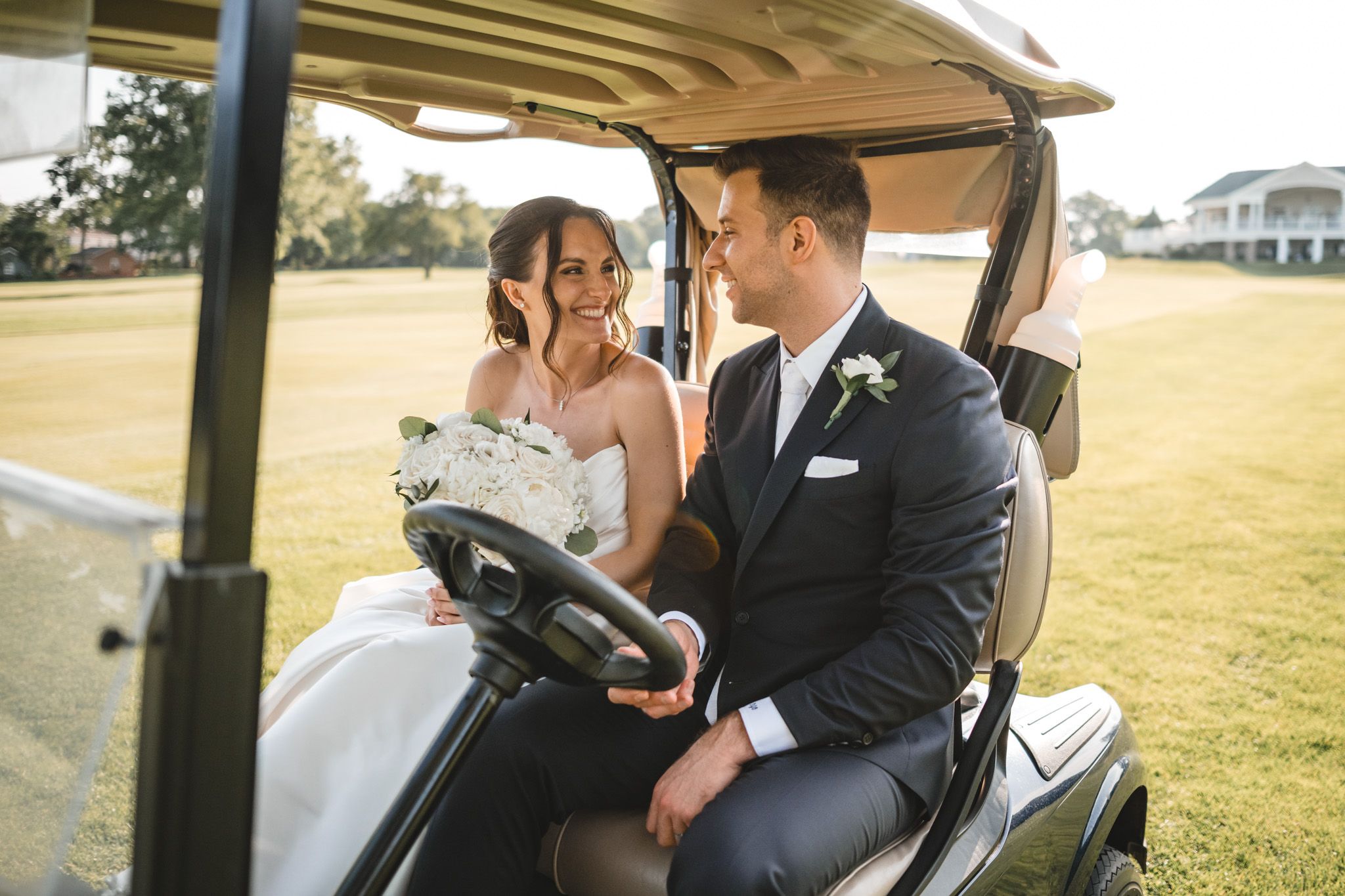 A wedding couple rides together in a golf cart across a sunny golf course.