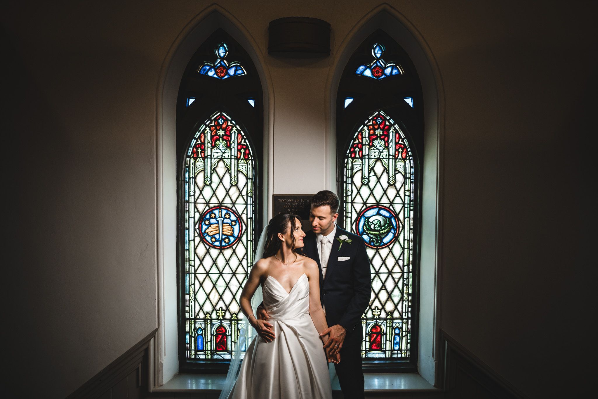 Stained glass windows cast dramatic light in a church interior during a wedding ceremony.