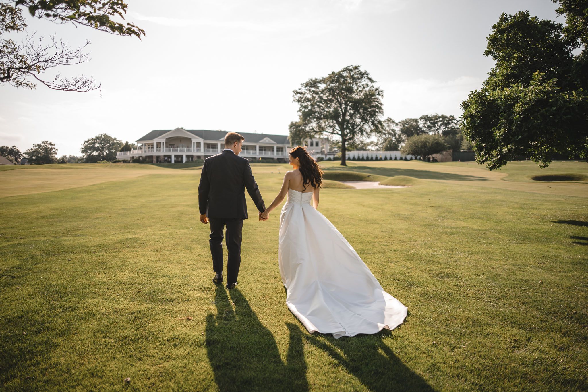 Wedding couple walks hand in hand across a sunlit grassy field on their special day.