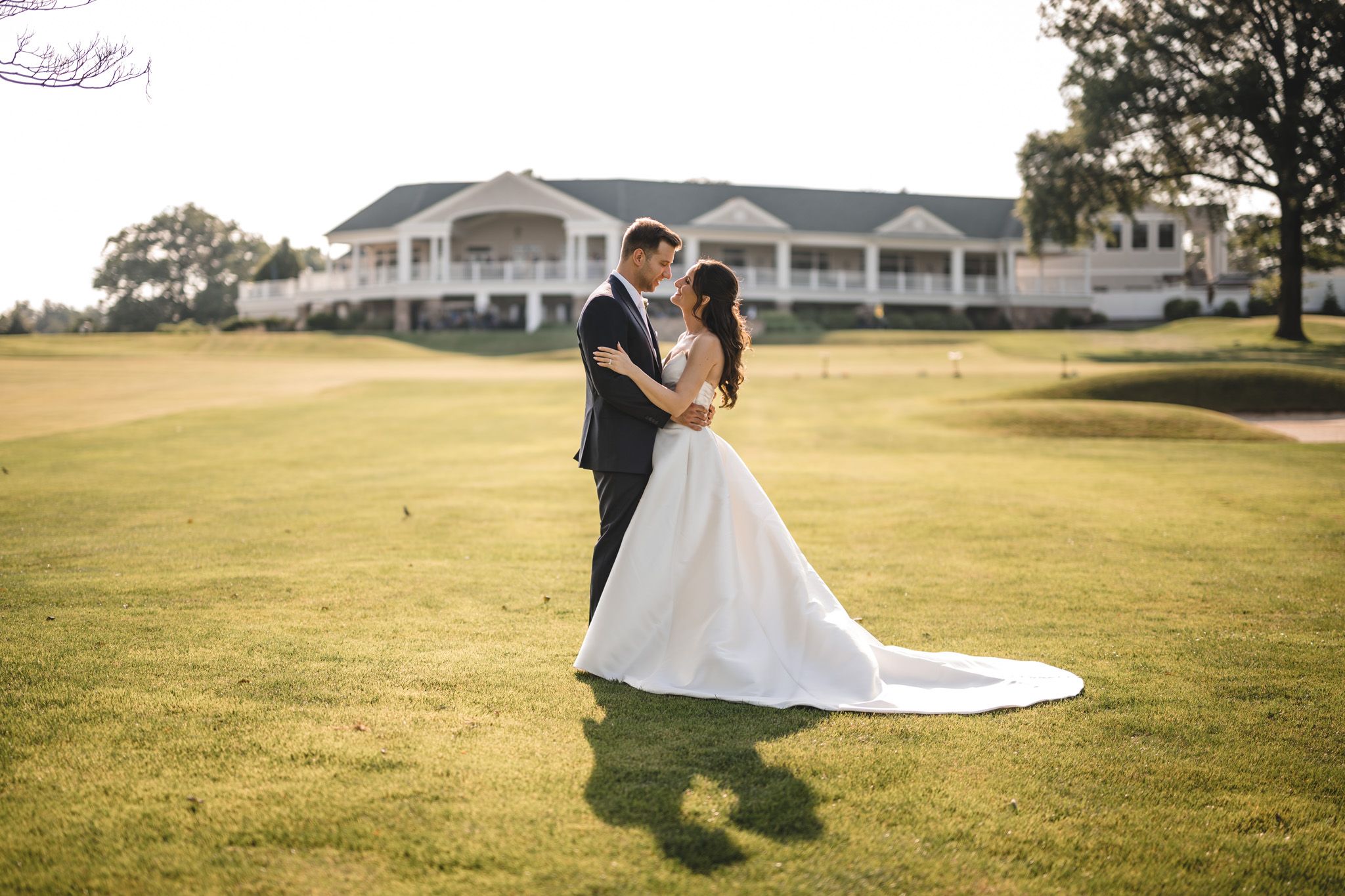 Couple in wedding attire strolls across a scenic golf course with colonial-style buildings in background.